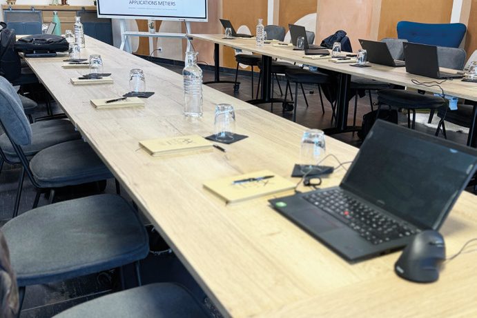 Grande table de travail avec ordinateurs et carnets, écran au fond de la salle