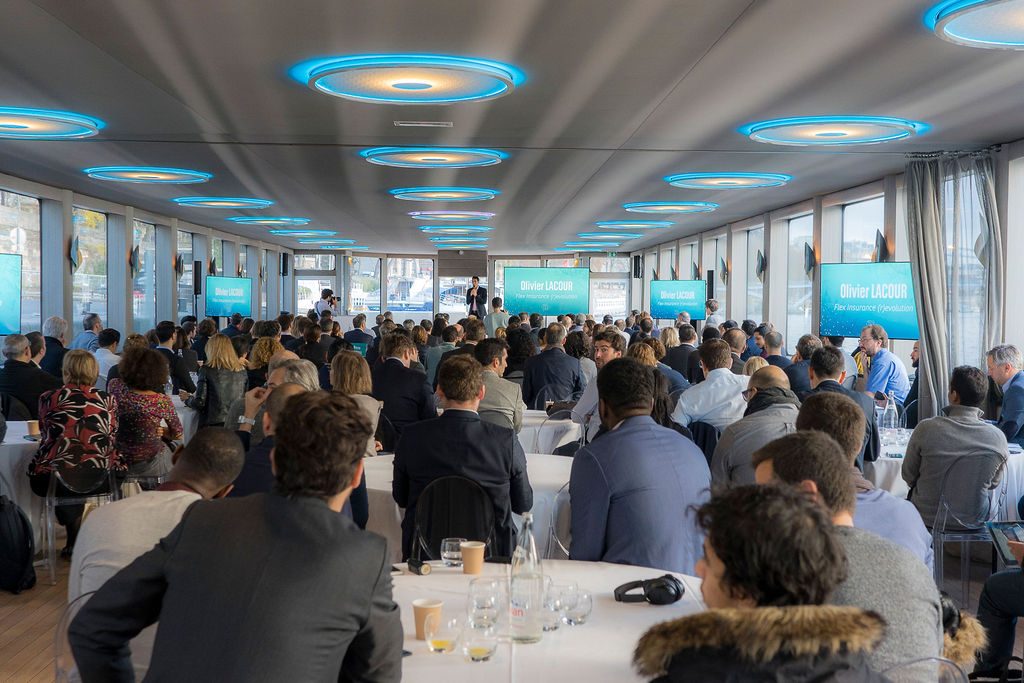 Une salle comble assise autour de tables rondes, face aux écrans d'une conférence à bord