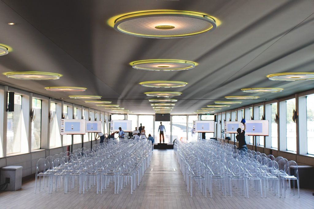 La grande salle installée en théâtre, chaises transparentes face à la scène et aux écrans