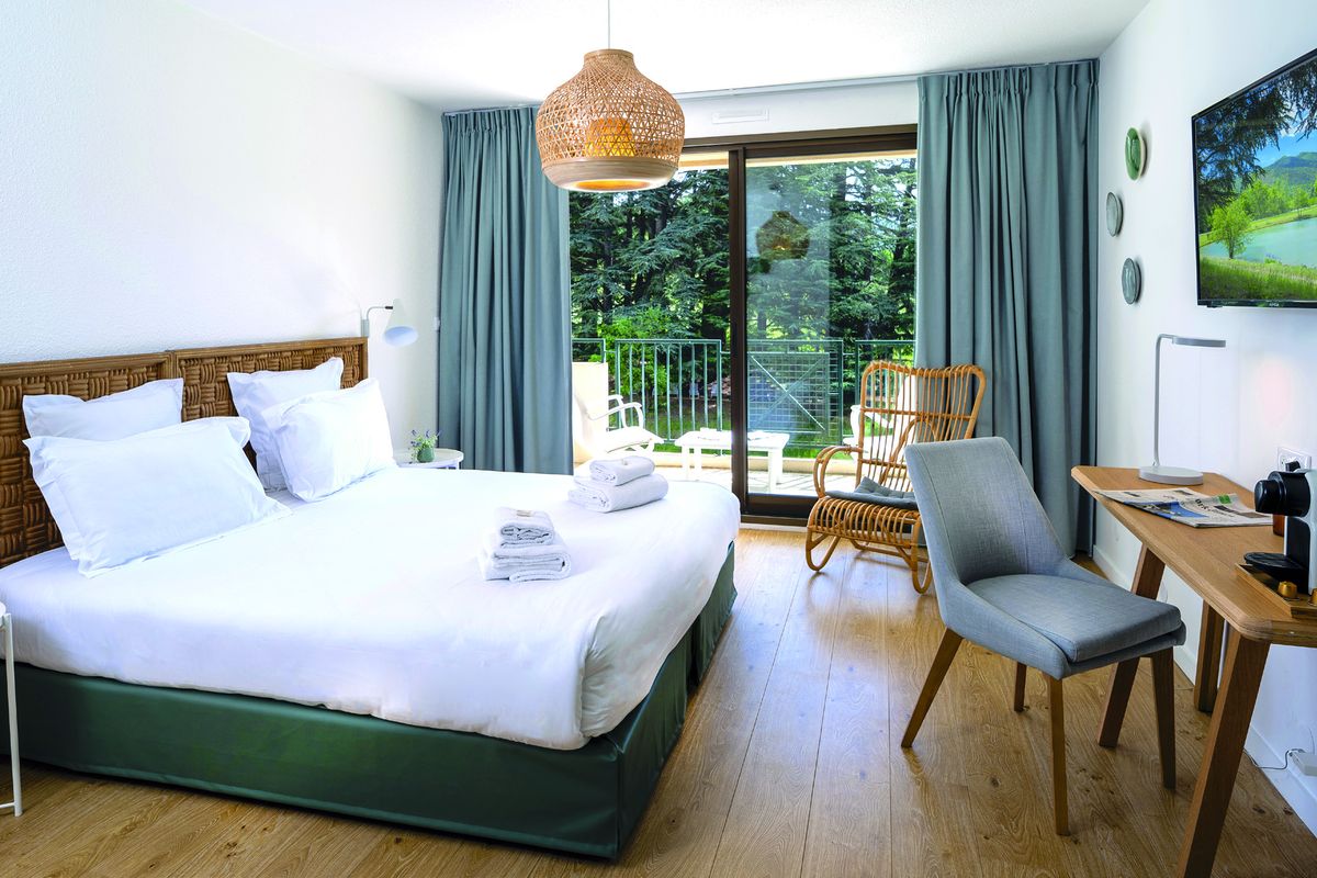 Chambre claire avec grand lit, bureau et baie vitrée ouverte sur la terrasse et la forêt